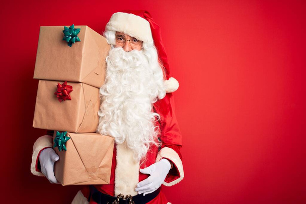 älterer Mann im Weihnachtsmann-Kostüm mit einem Turm von Geschenken vor isoliertem rotem Hintergrund, mit einem glücklichen Gesicht im Stehen und einem selbstbewussten Lächeln, das Zähne zeigt - Foto, Bild