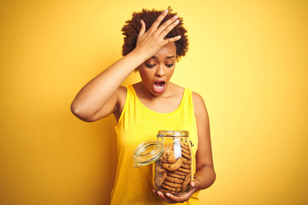 Femme afro-américaine tenant un pot de biscuits aux pépites de chocolat sur fond jaune stressé avec la main sur la tête, choqué par la honte et le visage surprise, en colère et frustré. Peur et contrariété par erreur
. - Photo, image