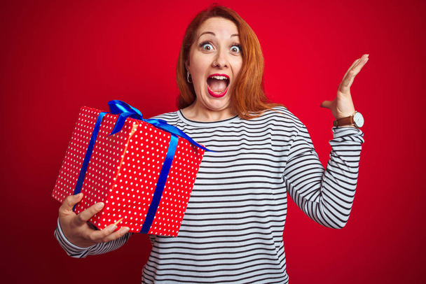 Young beautiful redhead woman holding birthday gift over red isolated background very happy and excited, winner expression celebrating victory screaming with big smile and raised hands - Photo, Image