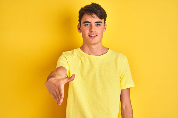 Tiener jongen draagt geel t-shirt over geïsoleerde achtergrond glimlachend vriendelijk aanbieden van handdruk als groet en gastvrij. Succesvolle bedrijfsvoering. - Foto, afbeelding