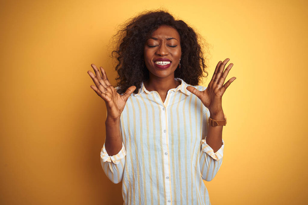African american woman wearing striped shirt standing over isolated yellow background celebrating mad and crazy for success with arms raised and closed eyes screaming excited. Winner concept - Foto, imagen