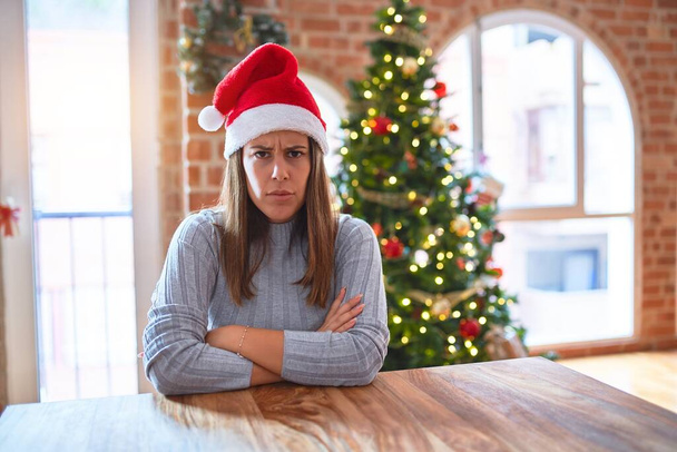 Jovem mulher bonita vestindo chapéu de Papai Noel na mesa em casa em torno de decoração de Natal cético e nervoso, desaprovando expressão no rosto com os braços cruzados. Pessoa negativa
. - Foto, Imagem
