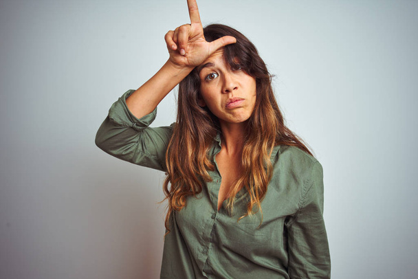 Young beautiful woman wearing green shirt standing over grey isolated background making fun of people with fingers on forehead doing loser gesture mocking and insulting. - Photo, Image