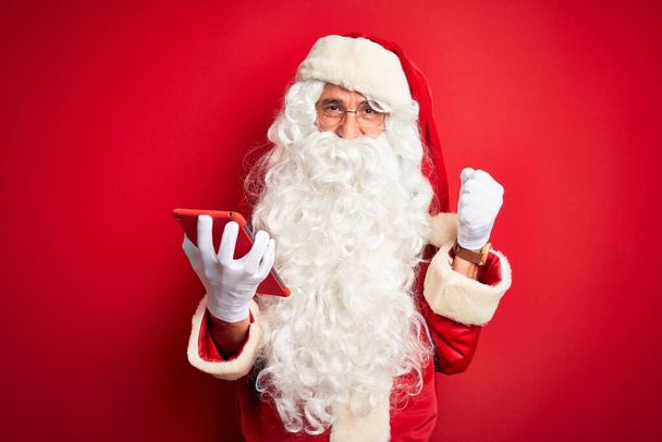 Middelbare leeftijd man dragen Santa Claus kostuum met behulp van tablet over geïsoleerde rode achtergrond schreeuwen trots en vieren overwinning en succes zeer opgewonden, juichen emotie - Foto, afbeelding
