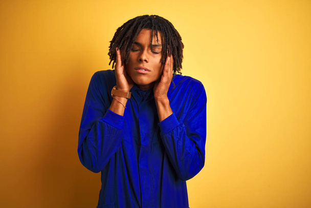 Afro worker man with dreadlocks wearing mechanic uniform over isolated yellow background with hand on head for pain in head because stress. Suffering migraine. - Foto, Imagen
