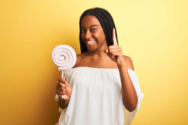 A fiatal afro-amerikai nő cukorkát eszik, elszigetelt sárga háttér felett áll meglepve egy ötlettel vagy kérdéssel, ami ujjal mutogat boldog arccal. - Fotó, kép