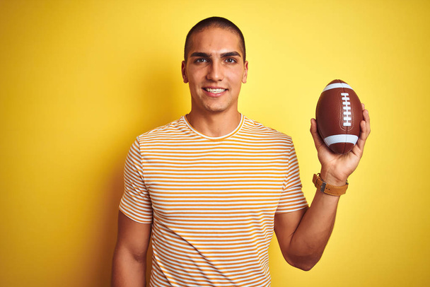 Молодий регбіст чоловік тримає футбольний м'яч над жовтим ізольованим тлом з щасливим обличчям, що стоїть і посміхається з впевненою посмішкою, показуючи зуби
 - Фото, зображення