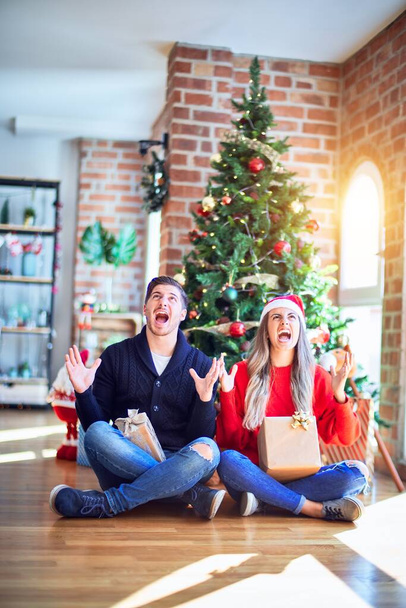 Jeune couple portant santa claus chapeau assis sur le sol autour de l'arbre de Noël à la maison fou et fou crier et crier avec l'expression agressive et les bras levés. Concept de frustration
. - Photo, image