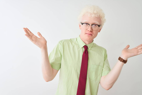 Joven hombre de negocios albino con camisa y corbata de pie sobre fondo blanco aislado expresión despistada y confusa con los brazos y las manos levantadas. Concepto de duda
. - Foto, Imagen