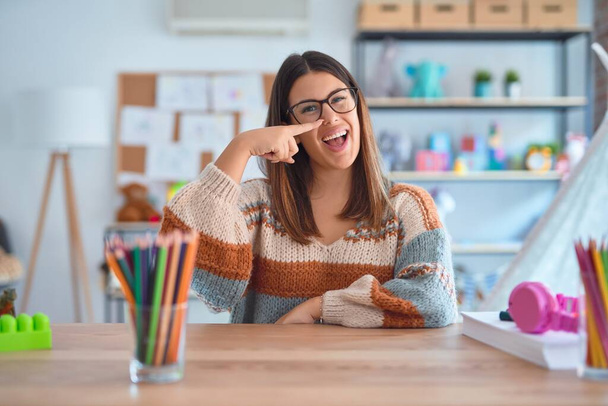 Young beautiful teacher woman wearing sweater and glasses sitting on desk at kindergarten Pointing with hand finger to face and nose, smiling cheerful. Beauty concept - Photo, Image