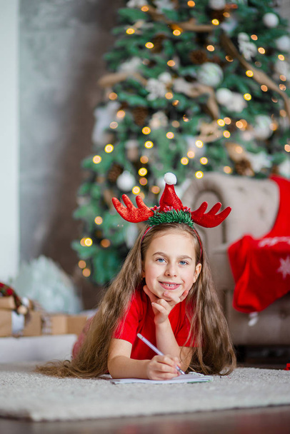 A cute girl in a Santa hat writes a letter to Santa near the Christmas tree. Happy childhood, a time for fulfilling desires. - Фото, изображение