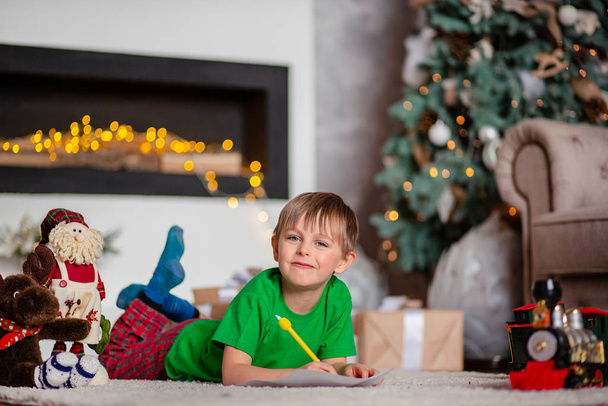 Vrolijke jongen is een brief aan de Kerstman, vlakbij de kerstboom. Gelukkige kindertijd, tijd voor het vervullen van wensen. - Foto, afbeelding