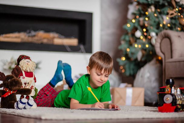 Cheerful boy is a letter to Santa, near the Christmas tree. Happy childhood, time for fulfilling wishes. - Foto, afbeelding