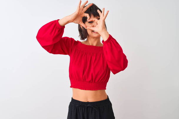 Młoda piękna kobieta w czerwonym letnim t-shircie stojąca nad odizolowanym białym tłem Robi kształt serca z dłoni i palców uśmiechając się patrząc przez znak - Zdjęcie, obraz
