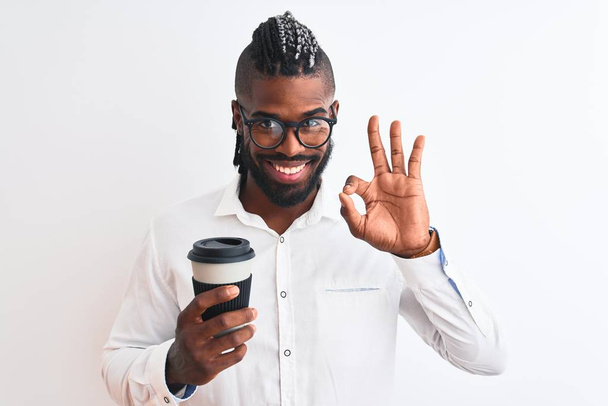 Afrykański amerykański biznesmen z warkoczami pijący kawę na białym tle robi znak ok palcami, doskonały symbol - Zdjęcie, obraz