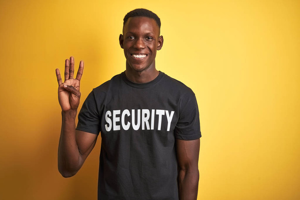 L'uomo afroamericano di salvaguardia indossa l'uniforme di sicurezza su sfondo giallo isolato mostrando e indicando con le dita il numero quattro mentre sorride fiducioso e felice
. - Foto, immagini