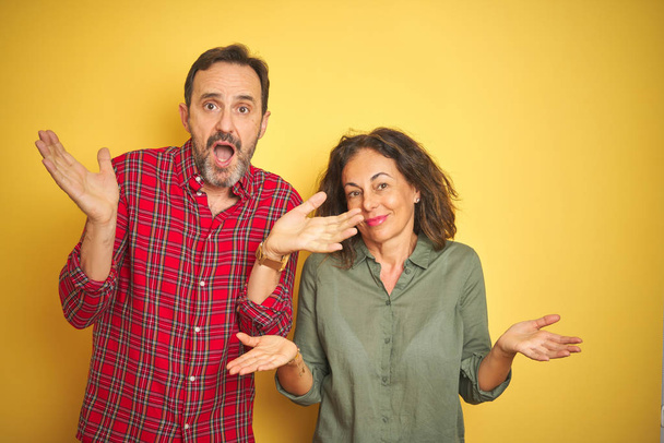 Beautiful middle age couple over isolated yellow background clueless and confused expression with arms and hands raised. Doubt concept. - Photo, Image