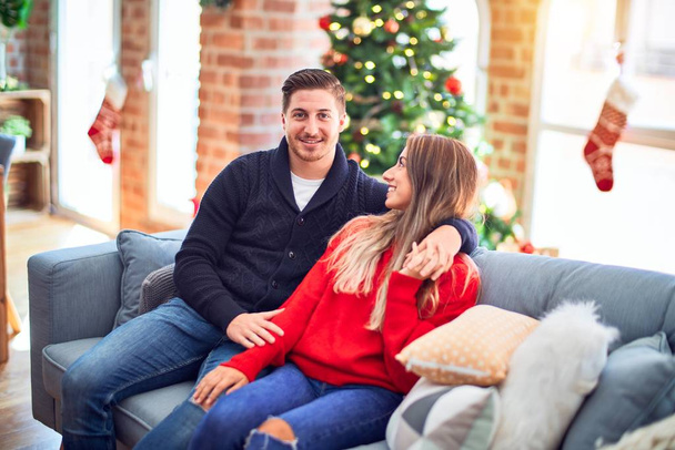 Young beautiful couple smiling happy and confident. Sitting on the sofa hugging around christmas tree at home - Фото, изображение