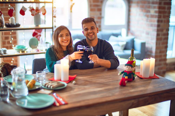 Young beautiful couple smiling happy and confident. Toasting with cup of wine celebrating christmas at home - Photo, Image