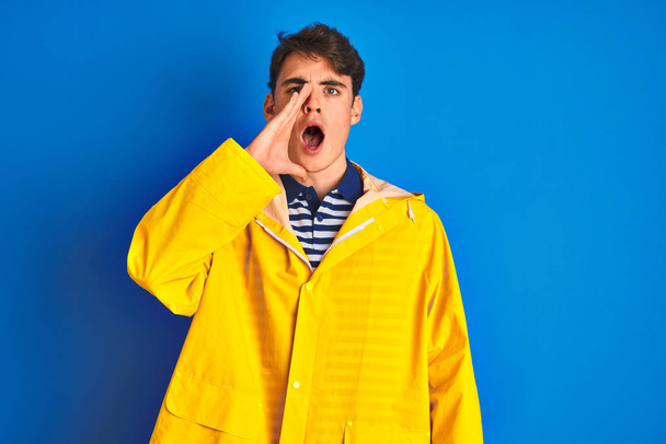 Έφηβος ψαράς που φορούσε κίτρινο αδιάβροχο πάνω από απομονωμένο φόντο φωνάζοντας και ουρλιάζοντας δυνατά στο πλάι με το χέρι στο στόμα. Έννοια επικοινωνίας. - Φωτογραφία, εικόνα
