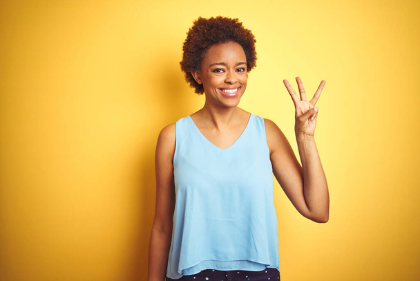 Gyönyörű afro-amerikai nő elegáns inget visel elszigetelt sárga háttér mutatja, és rámutatva ujjak száma 3, miközben mosolygós magabiztos és boldog. - Fotó, kép