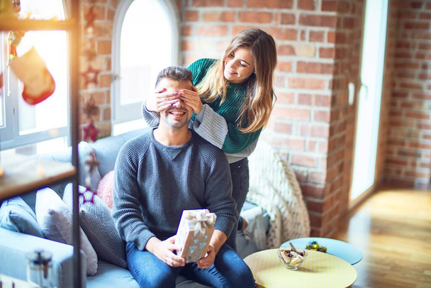 Молодая красивая пара улыбается счастливой и уверенной. Женщина, закрывающая глаза мужчины, окружает ее подарком вокруг рождественского украшения дома
 - Фото, изображение
