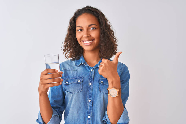 Молодая бразильская женщина держит стакан воды стоя на изолированном белом фоне счастливый с большой улыбкой делает хорошо знак, палец вверх пальцами, отличный знак
 - Фото, изображение