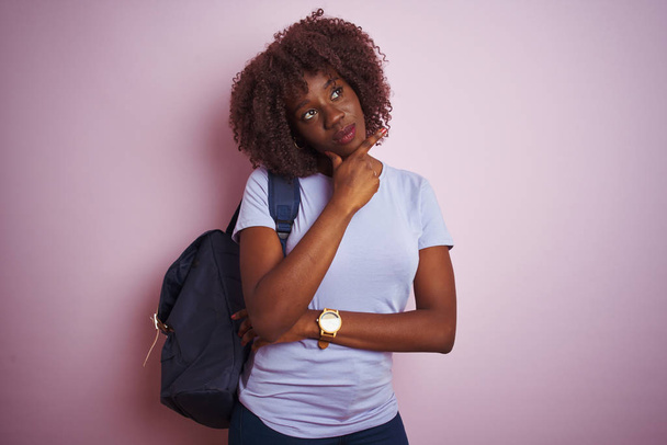 Молодая африканская туристка в рюкзаке, стоящая на изолированном розовом фоне с рукой на подбородке, думает о вопросе, задумчивом выражении лица. Улыбается с вдумчивым лицом. Концепция сомнений
. - Фото, изображение