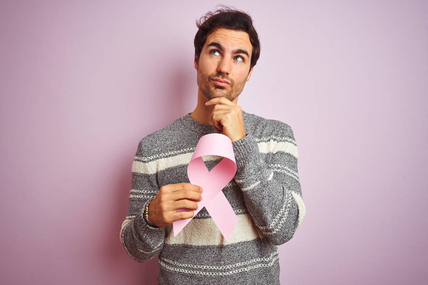 Młody przystojny mężczyzna trzymający wstążkę nowotworową stojącą nad odizolowanym różowym tłem poważna twarz myśląca o pytaniu, bardzo zdezorientowany pomysł - Zdjęcie, obraz