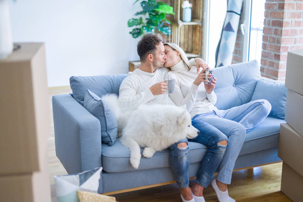 Young beautiful couple with dog sitting on the sofa drinking coffee at new home around cardboard boxes - Photo, Image