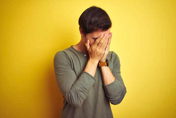 Молодой красивый мужчина в обычной футболке стоит на изолированном желтом фоне с грустным лицом, прикрывающим лицо руками во время плача. Концепция депрессии
. - Фото, изображение