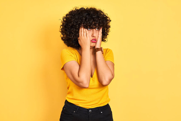 Young arab woman with curly hair wearing t-shirt standing over isolated yellow background Tired hands covering face, depression and sadness, upset and irritated for problem - 写真・画像