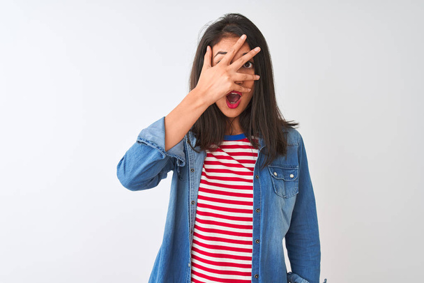 Young chinese woman wearing striped t-shirt and denim shirt over isolated white background peeking in shock covering face and eyes with hand, looking through fingers with embarrassed expression. - Photo, image