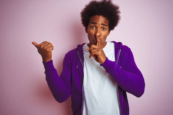 Jeune homme afro-américain portant un sweat-shirt violet debout sur un fond rose isolé demandant à être tranquille avec le doigt sur les lèvres pointant avec la main sur le côté. Silence et concept secret
. - Photo, image