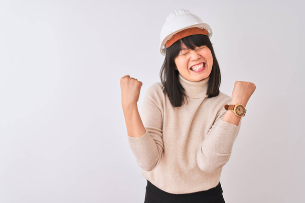 Young beautiful Chinese architect woman wearing helmet over isolated white background very happy and excited doing winner gesture with arms raised, smiling and screaming for success. Celebration concept. - Photo, Image