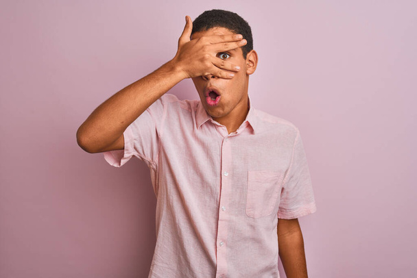 Mladý pohledný arabský muž v ležérní košili stojící nad izolovaným růžovým pozadím vykukující v šoku zakrývající obličej a oči rukou, dívající se skrz prsty s rozpačitým výrazem. - Fotografie, Obrázek