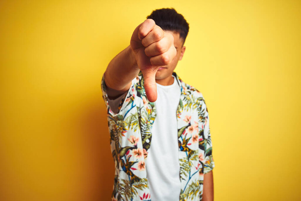 Young brazilian man on vacation wearing summer floral shirt over isolated yellow background looking unhappy and angry showing rejection and negative with thumbs down gesture. Bad expression. - Photo, image