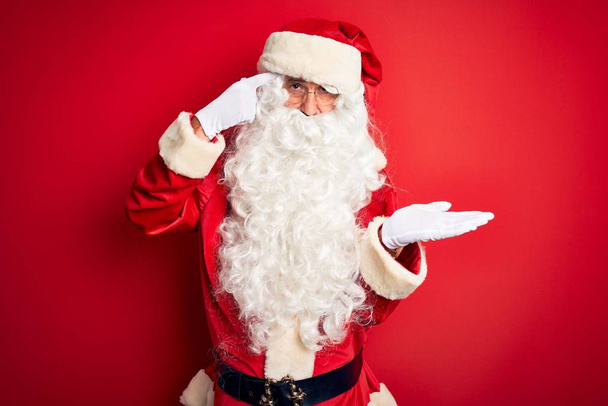 Homem bonito de meia idade vestindo traje de Papai Noel em pé sobre fundo vermelho isolado confuso e irritado com a palma aberta mostrando espaço de cópia e apontando o dedo para a testa. Pense nisso.
. - Foto, Imagem