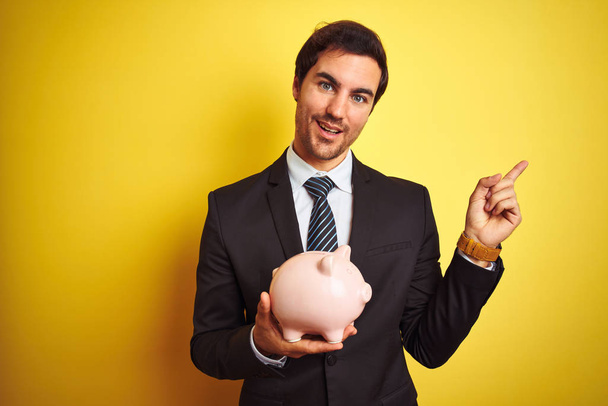 Young handsome businessman holding piggy bank over isolated yellow background very happy pointing with hand and finger to the side - Foto, Imagen