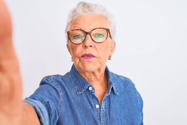 Седовласая женщина в джинсовой рубашке и очках делает селфи на изолированном белом фоне с уверенным выражением на умном лице, думая серьезно
 - Фото, изображение