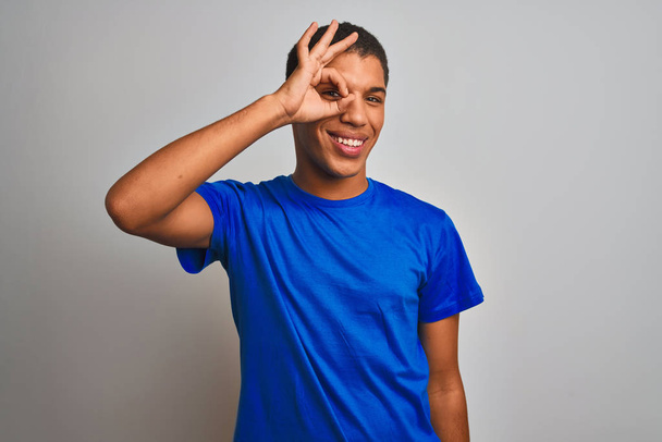 Young handsome arab man wearing blue t-shirt standing over isolated white background doing ok gesture with hand smiling, eye looking through fingers with happy face. - Foto, afbeelding