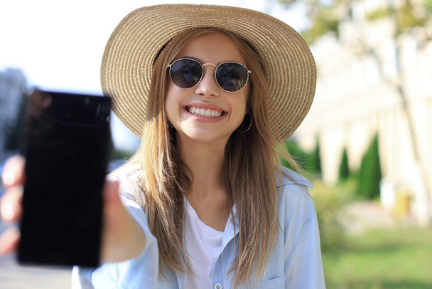 Ritratto di affascinante giovane donna che sorride e tiene lo smartphone, passeggiando per strada
. - Foto, immagini