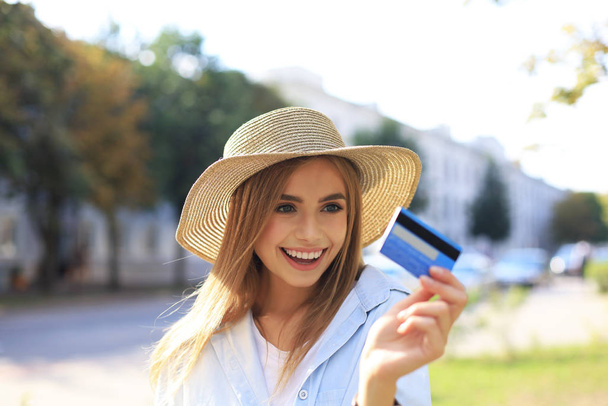 Cheerful young optimistic girl standing outdoors, holding credit card in hand. - Фото, изображение