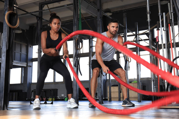 Athletic young couple with battle rope doing exercise in functional training fitness gym. - Фото, изображение