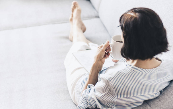 Middle-aged brunette woman on the gray sofa reading magazine wit - Zdjęcie, obraz