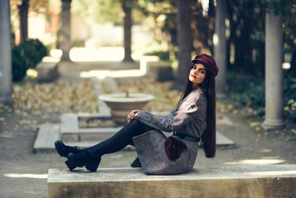 Young beautiful girl wearing winter coat and cap sitting on a bench in urban park. - Photo, Image