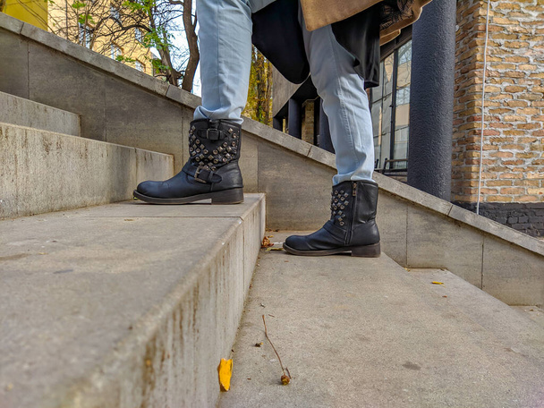Mulher indo para a frente um passo de cada vez escalando escadaria passo com botas punk pretas em passos de concreto. Outono outono, jeans azul e casaco amarelo
 - Foto, Imagem