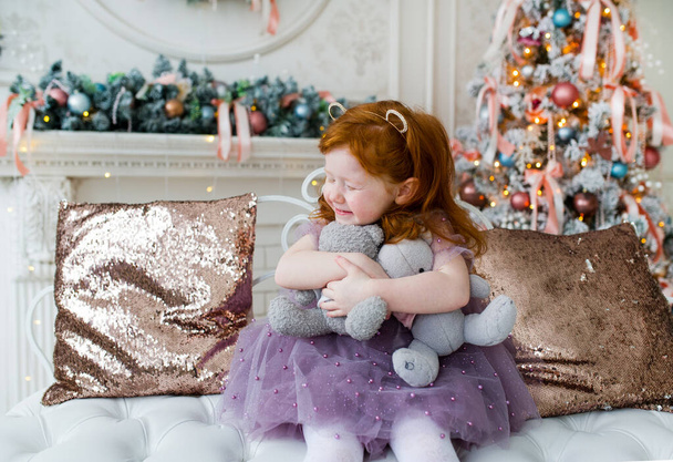 Niño pelirrojo con dos pequeños osos de peluche abrazan en la sala de estar decorada con accesorios de Navidad
. - Foto, Imagen