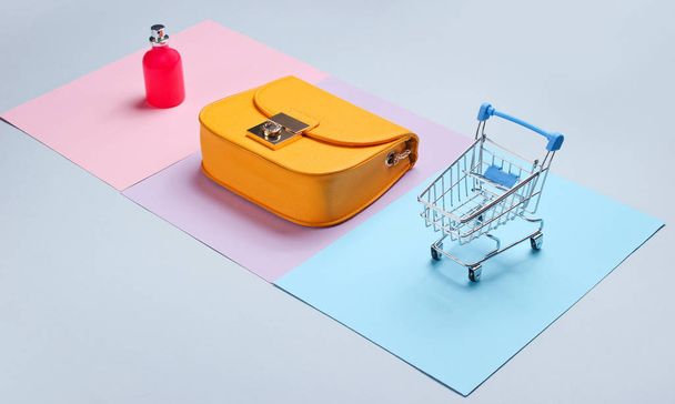 Магазинная минималистическая концепция. Желтая сумка, бутылка духов, мини-тележка для покупок на пастельном фоне. Вид сбоку
 - Фото, изображение