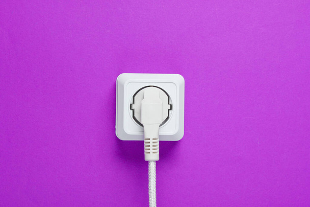 Cabo branco conectado à tomada de energia em um fundo de parede roxo
 - Foto, Imagem